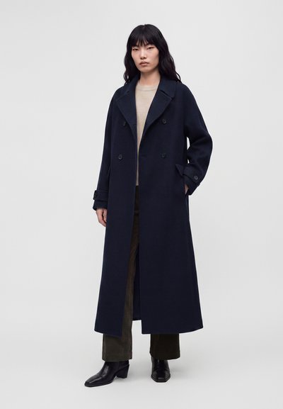 Jeune femme aux longs cheveux foncés portant un long manteau bleu marine à double boutonnage, un pull beige, un pantalon foncé et des bottines noires, debout devant un fond gris.
