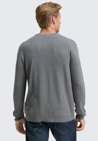 Man met blond haar in een effen grijze lange mouwen trui en donkerblauwe jeans, staand met zijn rug naar de camera op een effen lichte achtergrond.