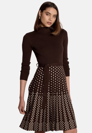 Frau trägt ein braunes, geripptes Langarmshirt und einen braunen Rock mit beigen Polka-Dots und einem passenden Stoffgürtel.