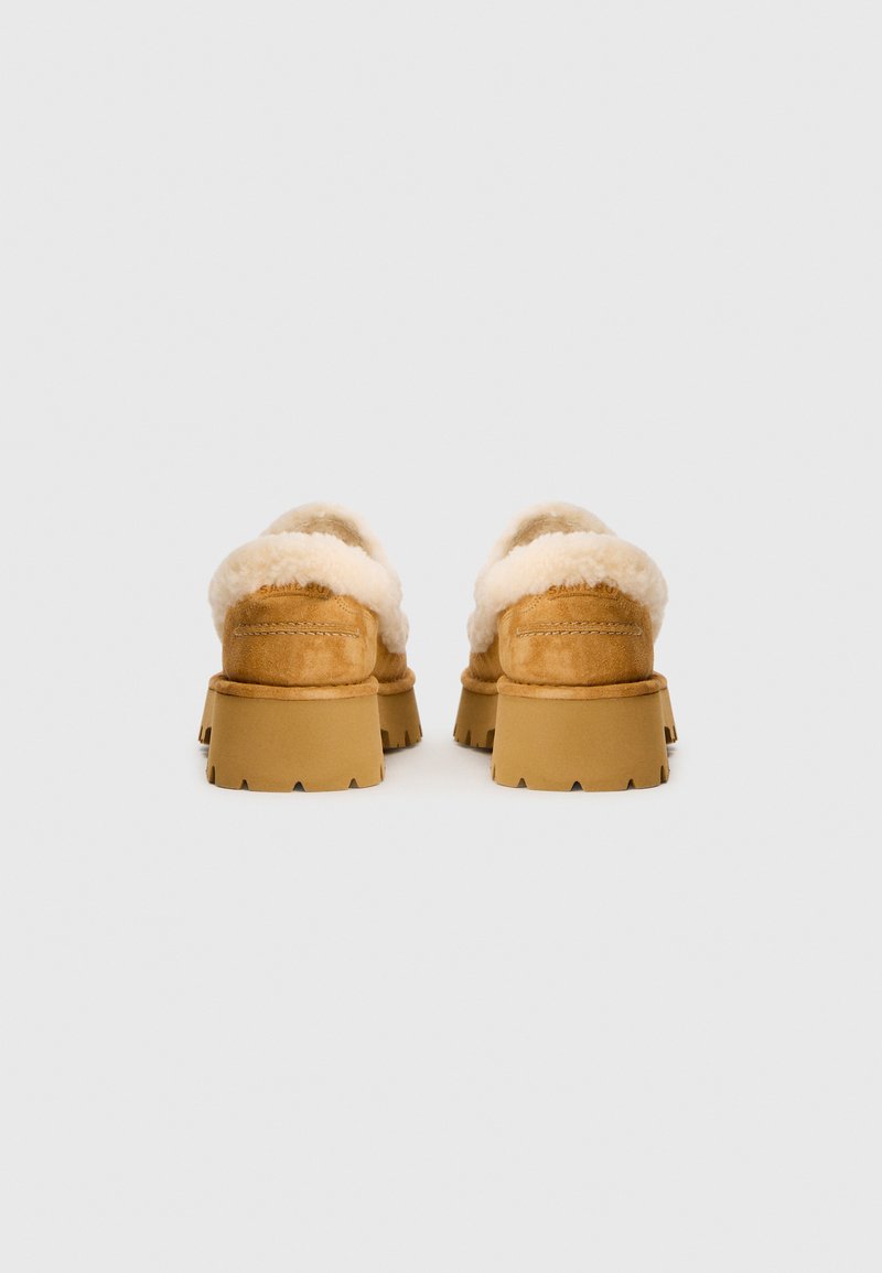 Suède instap schoenen met een witte fuzzy voering. Chunky tan rubberen zool met een getande rand, gezien vanaf de achterkant.