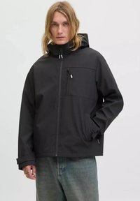 Eine blonde Person in einer schwarzen Kapuzenjacke mit Reißverschlusstaschen und blauen Jeans, die vor einem schlichten hellen Hintergrund steht.