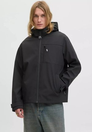 Blond person wearing a black hooded jacket with zippered pockets and blue jeans, standing against a plain light background.