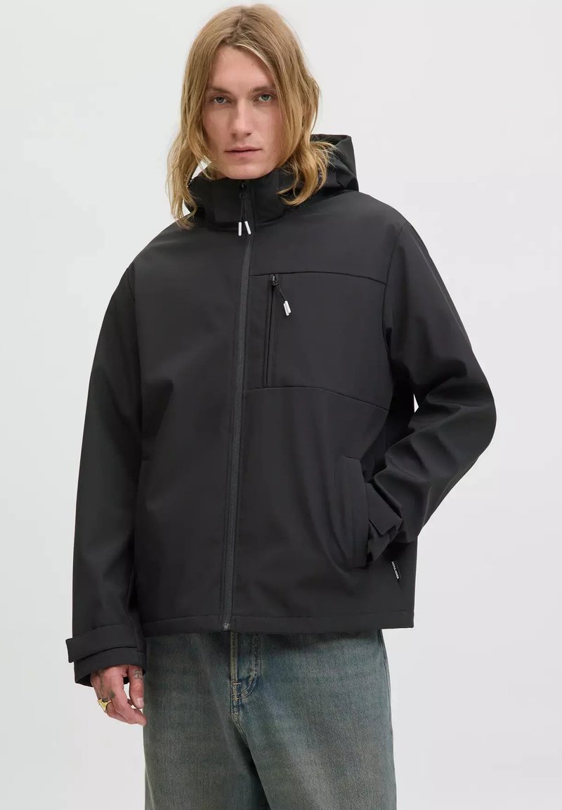 Eine blonde Person in einer schwarzen Kapuzenjacke mit Reißverschlusstaschen und blauen Jeans, die vor einem schlichten hellen Hintergrund steht.