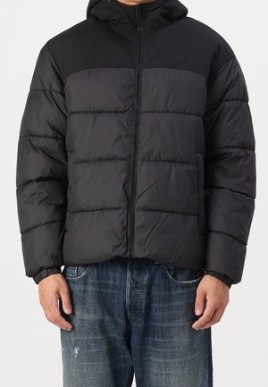 Homme portant une veste noire matelassée à capuche avec une fermeture éclair avant et un jean bleu, debout les mains détendues le long du corps.