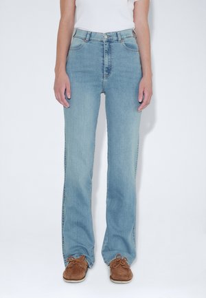 Mujer con vaqueros azul claro de pernera recta, camiseta blanca y zapatos casuales marrones, de pie sobre un fondo blanco liso.