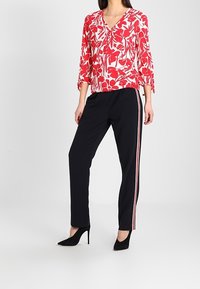 Rød blomstret bluse med V-hals og 3/4 lange ærmer, parret med sorte bukser med røde sidelister. Modellen bærer sorte stilletter.