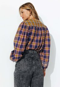 Femme aux cheveux blonds à épaule portant de grandes boucles d'oreilles en or, une blouse à carreaux moutarde et navy avec des manches bouffantes, et un jean noir taille haute.