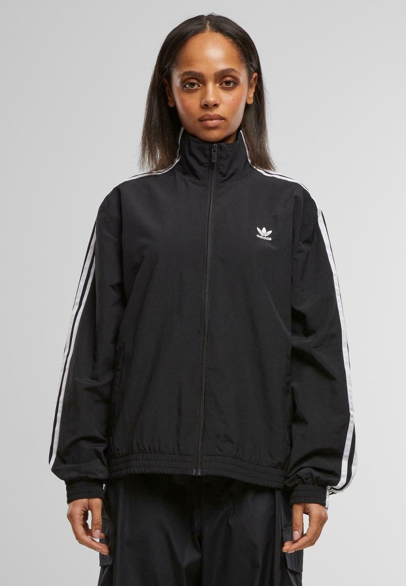 Schwarze Zip-Jacke mit hohem Kragen, weißen Seitenstreifen und einem gestickten Logo auf der Brust. Der Stoff wirkt glatt und leicht.