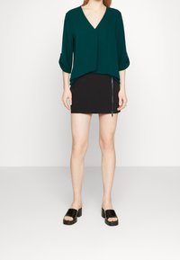 Blusa teal a V con maniche a tre quarti, abbinata a una mini gonna nera con zip laterale. Sandali neri chunky a scivolo.