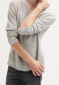 Maglione a maglia grigio chiaro, con maniche lunghe e vestibilità rilassata, realizzato in un tessuto testurizzato. Presenta un collo a V e maniche arrotolate, abbinato a pantaloni scuri.