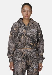Haut de hoodie zippé coupé en imprimé camouflage avec un mélange de bruns et de verts, doté d'une poche avant et d'un logo graphique sur la poitrine.