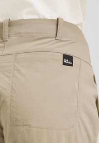 Pantaloni in tessuto beige con una tasca posteriore dotata di un'etichetta nera con la scritta "Jack Wolfskin". La texture sembra liscia con cuciture visibili.
