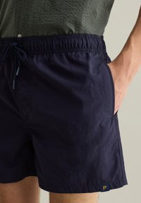 Pantaloni da bagno blu navy con vita elastica, coulisse, tasche laterali e un piccolo patch giallo sul bordo. Tessuto leggero.