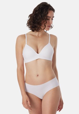 Femme portant un soutien-gorge blanc sans coutures et un sous-vêtement taille basse assorti, regardant vers le bas sur sa droite, sur un fond clair uni.