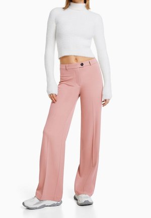 Roze wijdvallende broek met voorplooien, hoge taille en knoopsluiting, gecombineerd met een cropped witte textuursweater en grijze sneakers.