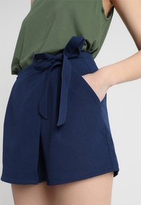 Tmavě modré šortky z texturované látky, s vázáním v pase, bočními kapsami a střihem na míru pro přiléhavý vzhled.