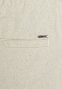 Beige fabric bag with textured surface, featuring a small metallic brand tag and a zippered pocket for secure storage.