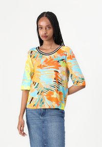 Buntes Kurzarmoberteil mit einem tropischen Muster in Blau, Gelb und Orange. Rundhalsausschnitt mit schwarzem Saum. Leichtes Material, lockere Passform.