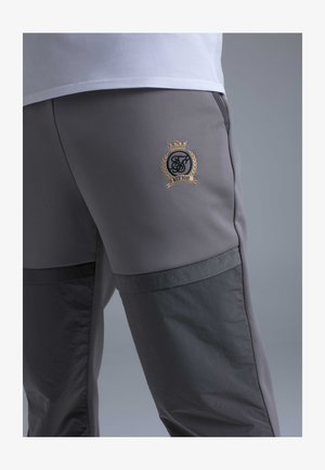 Nahaufnahme von grauen Hosen mit gesteppten dunkelgrauen Einsätzen und einem goldbestickten Wappen mit Krone, Kranz und dem Schriftzug "EST 2012".