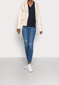 Beige Kunstleder-Motorradjacke über einem navy V-Ausschnitt-Top, kombiniert mit ausgewaschenen blauen Skinny-Jeans und weißen Sneakers.