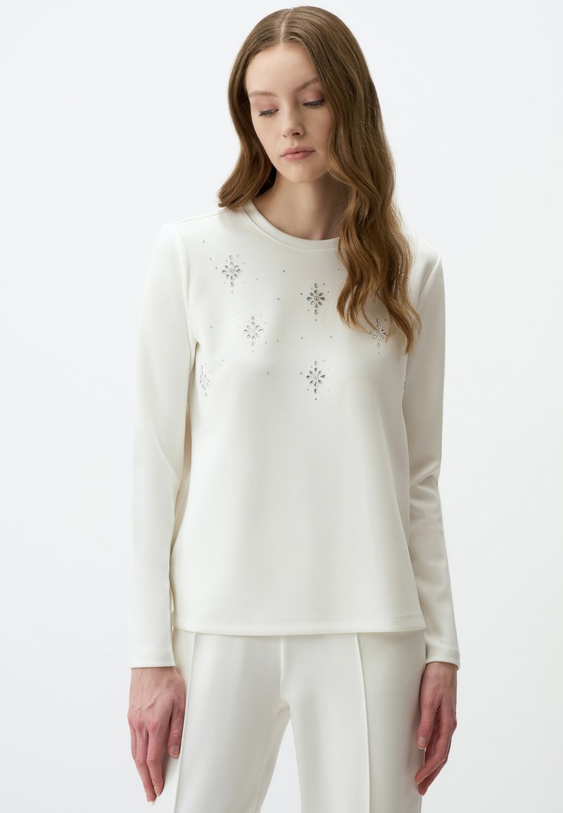 Witte longsleeve top met decoratieve zilveren versieringen in bloemmotieven. Gemaakt van gladde stof met een ronde hals.