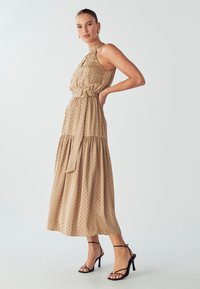 Hnedé bezrukávové šaty s bodkami, opasok v oblasti pása, sukňa s volánmi a hladký materiál. V kombinácii s čiernymi sandálmi na vysokých ihlových podpätkoch.