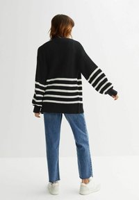 Person som står med ryggen mot kameran, iklädd en svart oversized tröja med vita ränder och blå jeans, framför en enkel vit bakgrund.