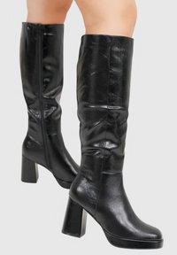 Bottes noires montantes en matériau brillant, avec un talon épais et une texture lisse, dotées d'une fermeture éclair sur le côté pour un enfilage facile.
