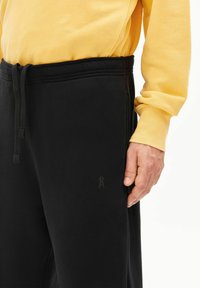 Zwarte joggingbroek met een zachte textuur, voorzien van een taillekoord en een klein logodetail op het been. Gecombineerd met een gele longsleeve.