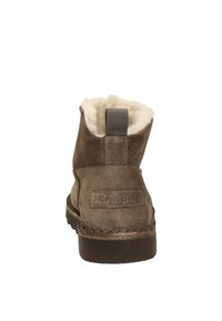 Braune Wildleder-Stiefeletten mit einem flauschigen weißen Futter, einer Zuglasche hinten und einer strukturierten Gummisohle. Branding an der Seite sichtbar.