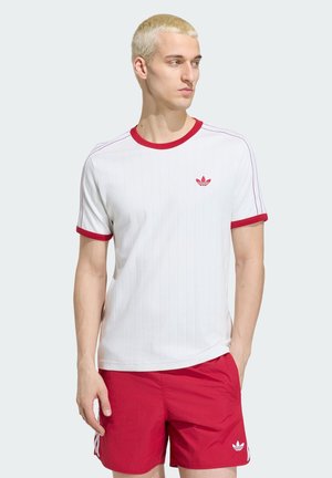 Jeune homme aux cheveux blonds courts portant un t-shirt blanc avec bordure rouge et logo, assorti à un short rouge avec logo blanc, debout devant un fond uni.