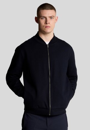 Sweatshirt zippé bleu marine avec col rond, poignets et ourlet côtelés. Tissu lisse avec un design minimaliste sans motifs ni logos apparents.
