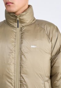 Pogled od blizu na osebo, ki nosi zadrgo bež pufast plašč z visokim ovratnikom in belim logotipom "OBEY" na prsih.