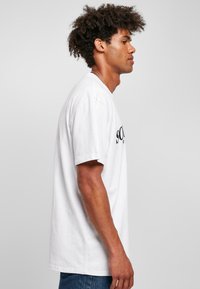 Camiseta de algodón blanca de manga corta, colgando de manera suelta. Texto negro en la parte frontal. Diseño simple, cuello redondo, corte recto. Pantalones vaqueros de denim azul.