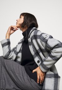 Manteau en laine à carreaux gris avec des accents noirs, porté par-dessus un col roulé noir à côtes. Le modèle pose assis avec un pantalon à motifs superposés et des cheveux courts.