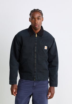 Uomo vestito con giacca zip blu scuro con colletto nero e pantaloni blu, in piedi contro uno sfondo chiaro e uniforme.