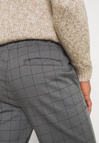 Šedé kostkované kalhoty s detailem zadní kapsy, zkombinované s texturovaným béžovým svetrem, zobrazující detailní pohled na látku a design.