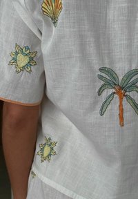 Baltas lino drabužis su ryškia išsiuvinėjimu, vaizduojančiu jūros kriaukles, augalus ir gėles mėlynos, oranžinės ir žalios spalvų, kontrastingomis siūlėmis.