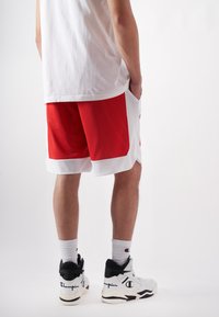 Rot-weiße Netz-Sportshorts mit seitlichen Taschen, kombiniert mit weißen Sneakers mit schwarzen Akzenten und einem Champion-Logo.