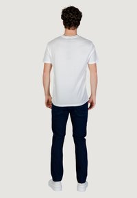 Homme aux cheveux bouclés portant un t-shirt blanc uni, un jean bleu foncé et des baskets blanches, debout de dos sur un fond neutre.
