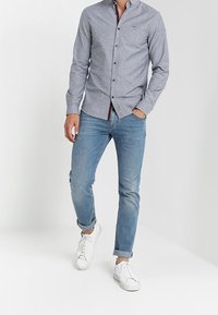 Mężczyzna w szarej koszuli na guziki, niebieskich podwiniętych dżinsach i białych trampkach, stojący na tle jasnego, jednolitego tła.