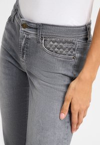 Graue Jeans aus Denim mit einer dekorativen Tasche im Diamantmuster und metallischen Akzenten. Der Stoff hat eine leicht strukturierte Oberfläche.