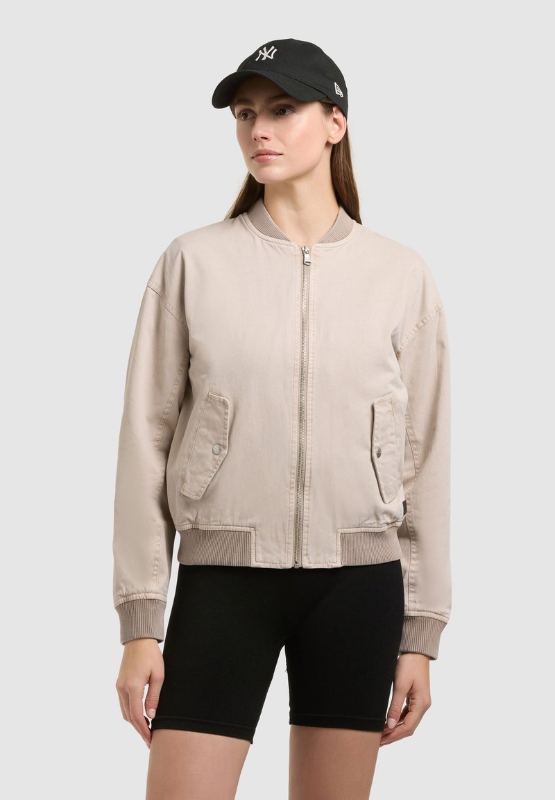 Femme portant une veste bomber beige zippée, un short de cyclisme noir et une casquette de baseball noire sur un fond gris uni.