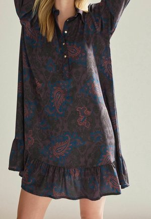 Femme portant une robe sombre avec des motifs cachemire bleus et marron et un ourlet à volants, debout, les bras levés sur un fond clair.