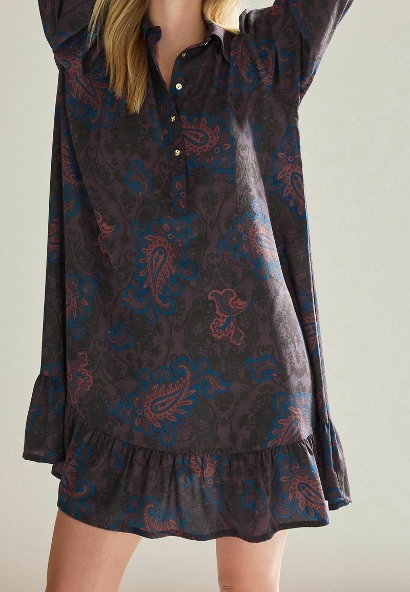 Femme portant une robe sombre avec des motifs cachemire bleus et marron et un ourlet à volants, debout, les bras levés sur un fond clair.