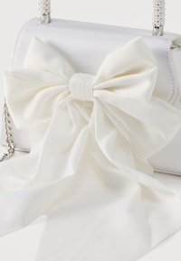 Λευκή σατέν τσάντα χειρός με λαβή διακοσμημένη με πέρλες και μεγάλο διακοσμητικό λευκό υφασμάτινο φιόγκο με ρέουσες λεπτομέρειες σε λευκή επιφάνεια.