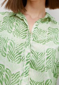 Blusa verde chiaro con motivo a foglia, realizzata in tessuto morbido con colletto e dettagli con bottoni lungo il davanti.