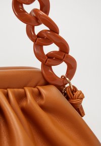 Borsa rotonda di colore arancio bruciato con finitura in pelle testurizzata e grande tracolla a catena lucida e intrecciata. Presenta una parte superiore elastica arricciata e un accento annodato.