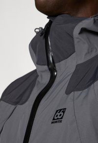 Chaqueta impermeable gris con cuello alto, cierre de cremallera y paneles oscuros contrastantes en los hombros. Logo bordado en el lado izquierdo del pecho.