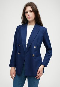 Marineblauwe dubbelbreasted blazer met gouden knopen, scherpe revers, voorzakken en een gladde textuur, gedragen over een witte top.